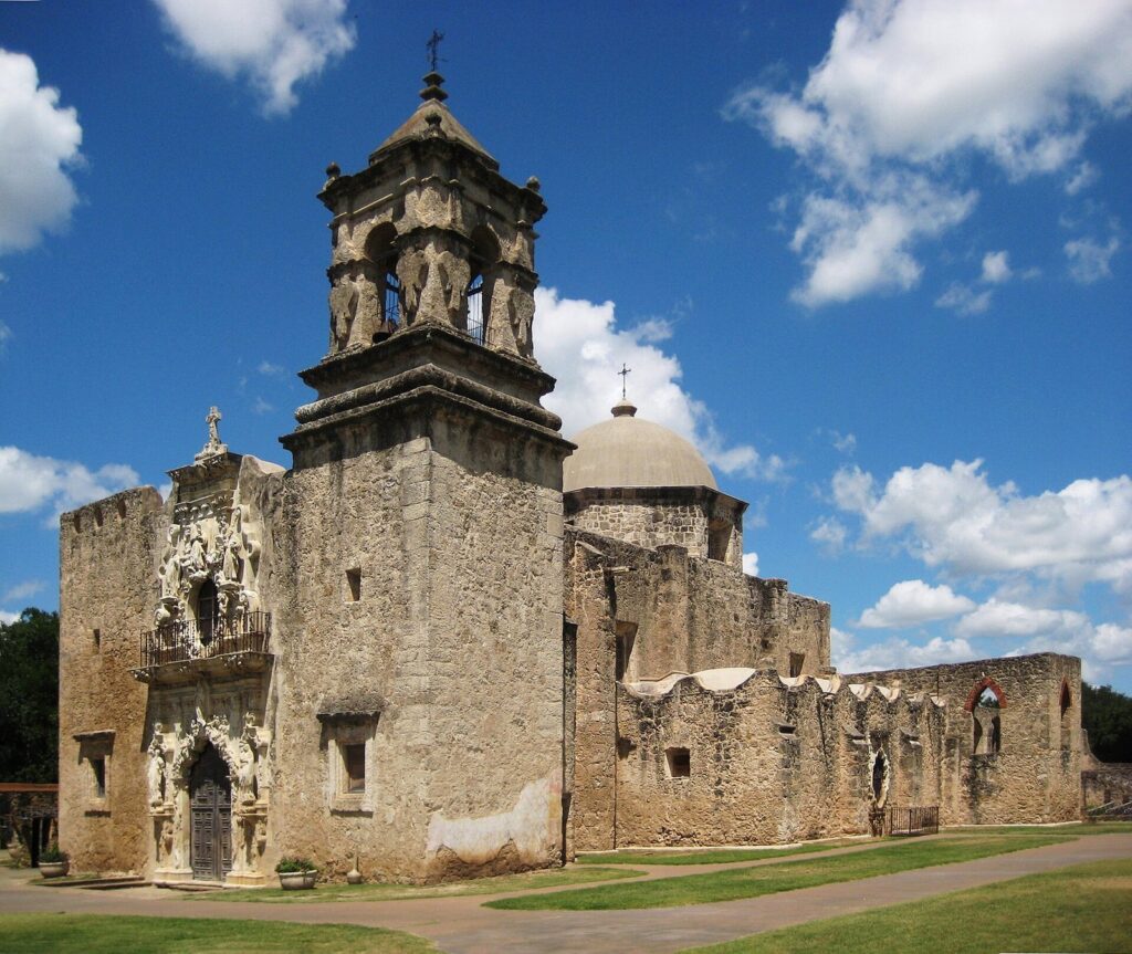 The limestone church and bell tower of Mission San José in San Antonio under a blue sky with scattered clouds
