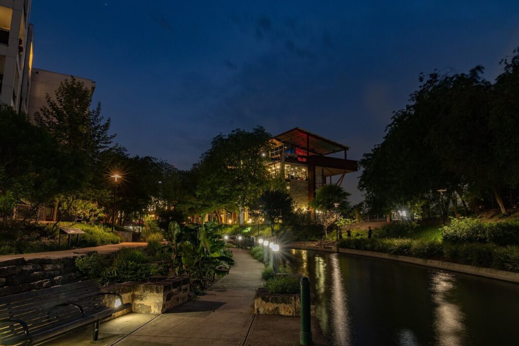 Path lights glowing along the Museum Reach of the San Antonio River Walk at dusk, with trees and a lit building beyond