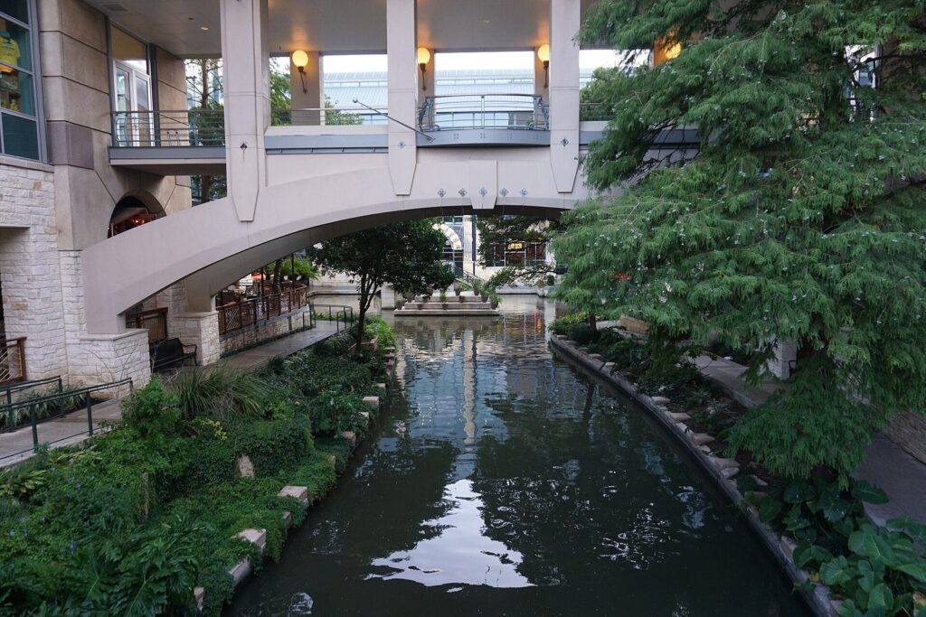 The River Walk channel curving under a covered walkway at the Shops at Rivercenter, with planted banks and river-level paths on both sides