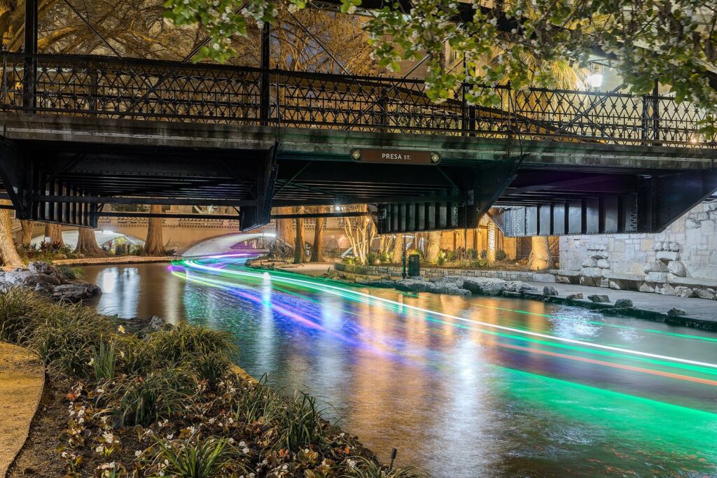 The San Antonio River Walk passing under the iron Presa Street bridge at dusk, with a boat's lights streaking across the water