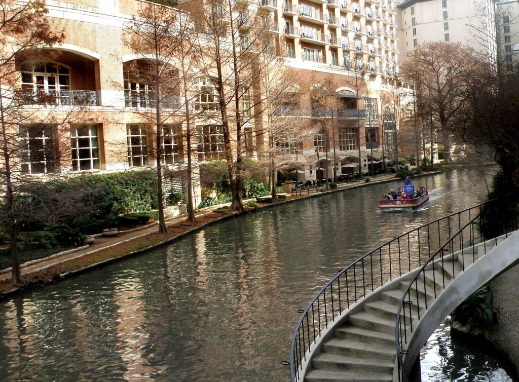 A curving stone stairway with a metal handrail leading down to the San Antonio River Walk beside cypress trees and a hotel