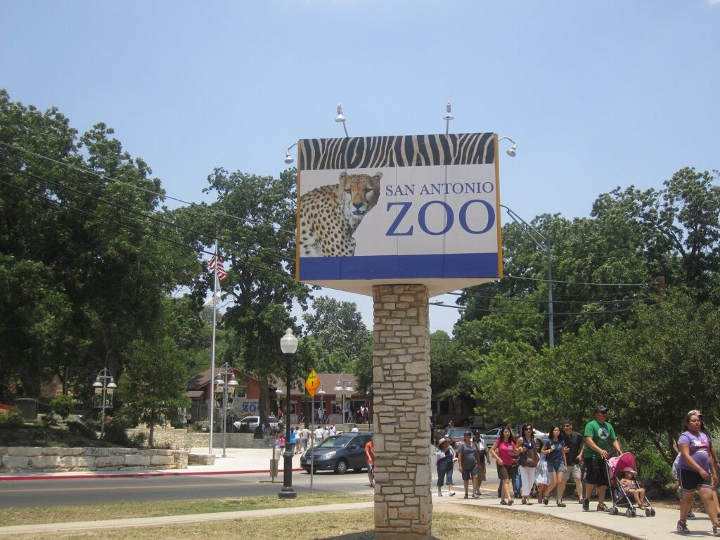 Families with a stroller walking past the San Antonio Zoo entrance sign under shade trees on a sunny day