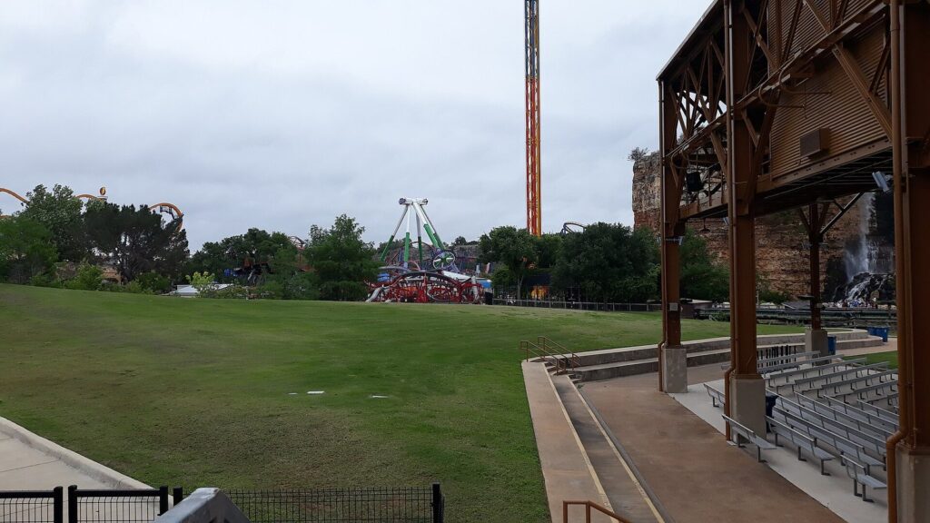 A wide lawn and covered seating inside Six Flags Fiesta Texas, with ride towers and the old quarry cliff in the distance under a cloudy sky
