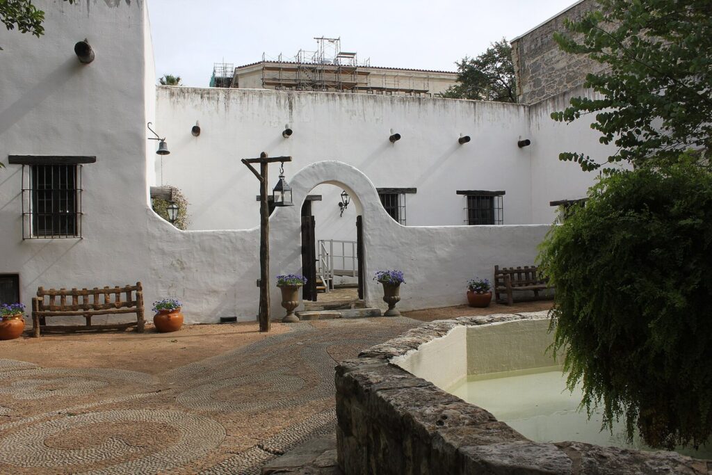 The whitewashed courtyard of the Spanish Governor's Palace in San Antonio, with an arched gate, wooden benches, potted plants and a stone fountain